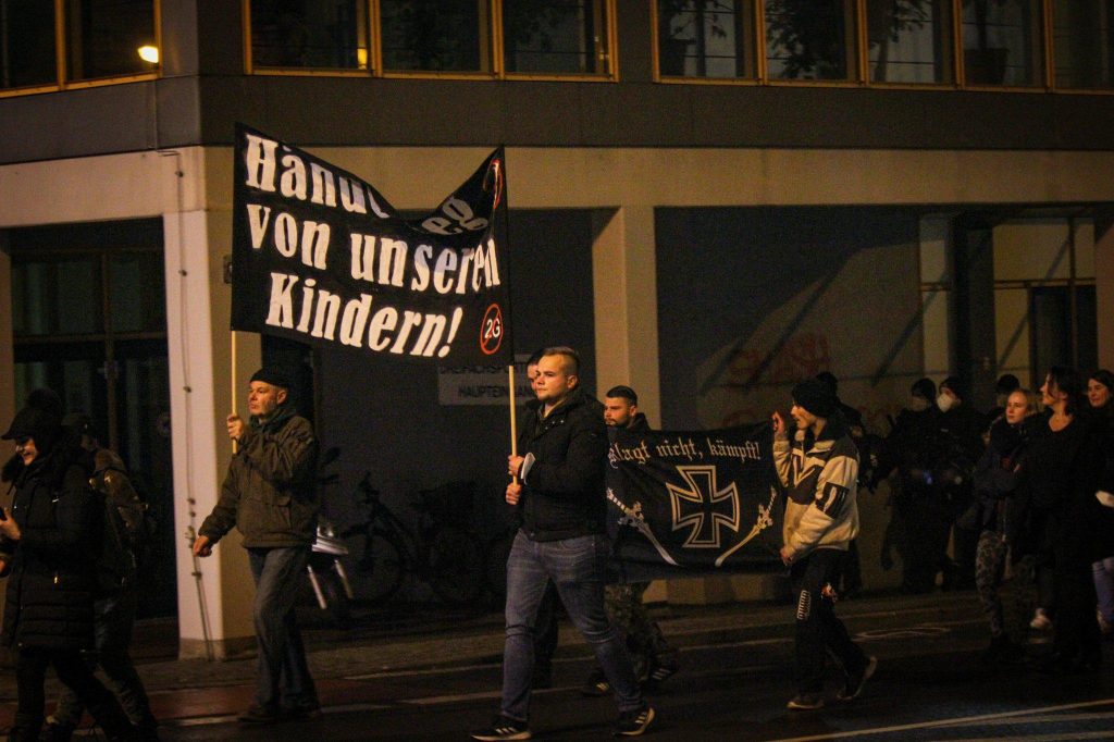 Roger Kuchenreuther (links) und Andreas Groh (rechts) tragen ein Banner mit der Aufschrift „Hände weg von unseren Kindern“. Hinter ihnen laufen zwei weitere Aktivisten mit einem Banner mit der Aufschrift „Klagt nicht, kämpft!“, auf dem ein Eisernes Kreuz und zwei Schwerter abgebildet sind.